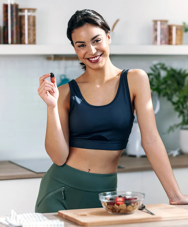 Woman in a kitchen with a bowl of fresh fruit, smiling while eating a berry